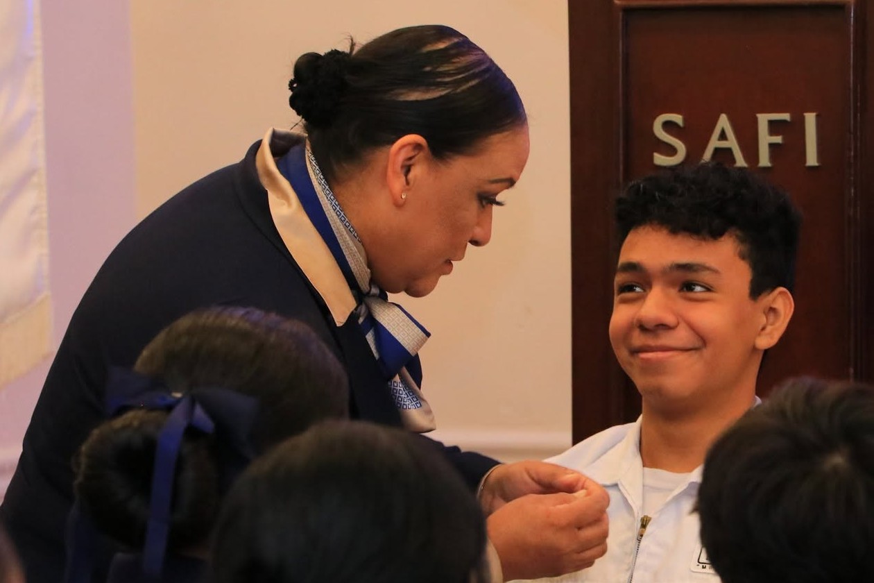 Una maestra coloca el prendedor a un alumno de Enfermería con uniforme blanco, que la mira sonriente