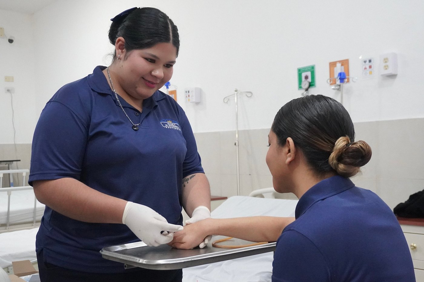 Alumna de Enfermería atiende con una sonrisa a una compañera durante la práctica